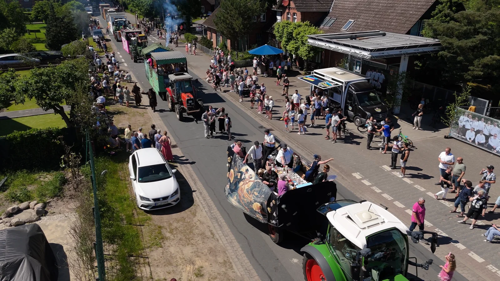 Drohnenaufnahme eines Festumzugs mit Traktoren, Festwagen und Gästen im Dorf